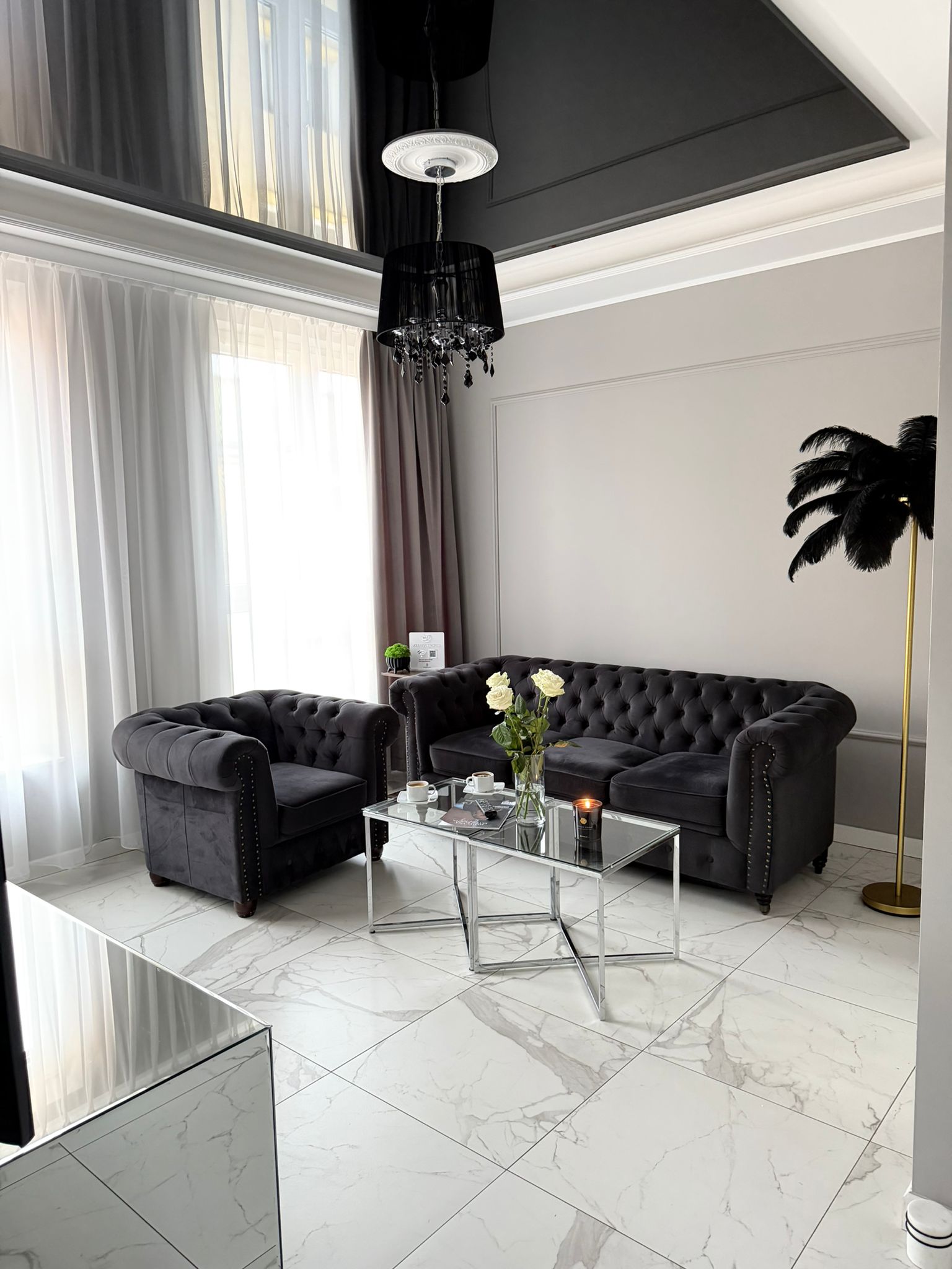 Salon z grafitową pikowaną sofą i fotelem, szklany stolik z białymi różami, marmurowa podłoga i czarny żyrandol