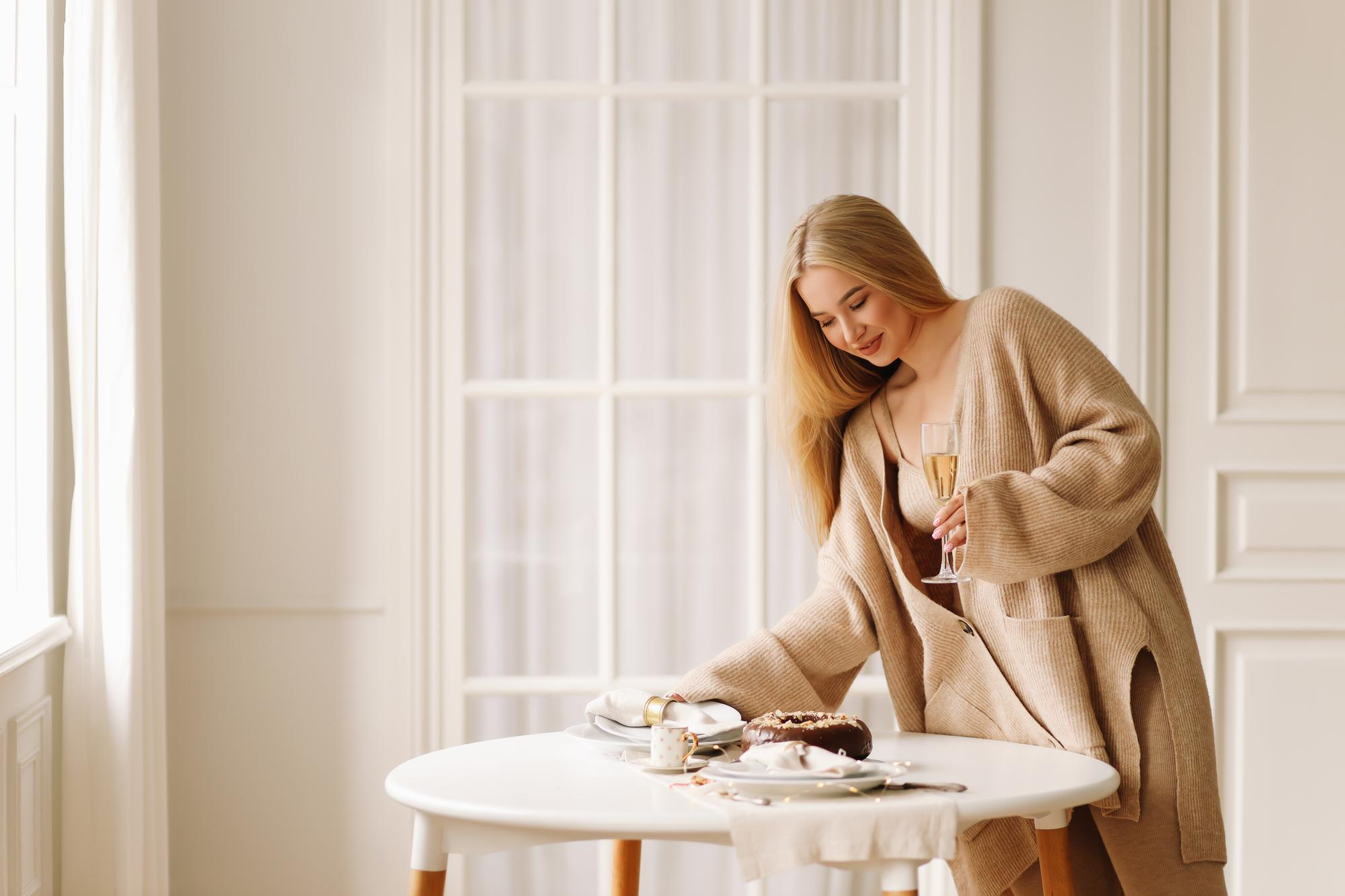 Blondynka w beżowym kardiganie nakrywa stół trzymając kieliszek szampana, obok czekoladowe ciasto