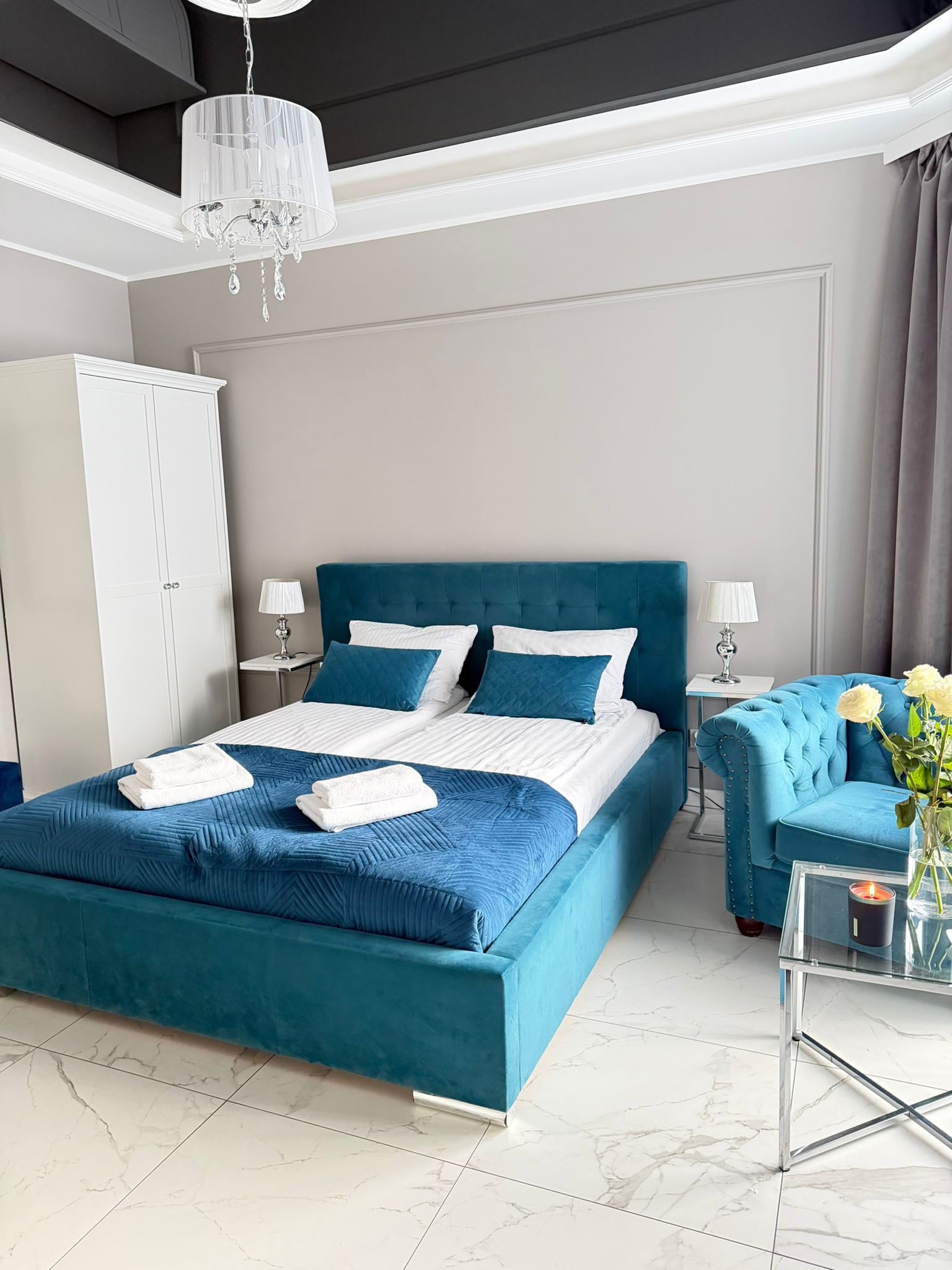Sypialnia z turkusowym aksamitnym łóżkiem i sofą, białą pościelą, złożonymi ręcznikami, żyrandolem i kwiatami